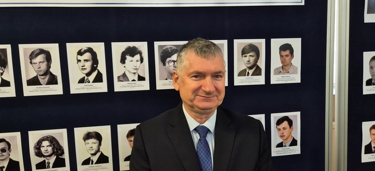 Dr hab. inż. Mariusz Korkosz, prof. uczelni na tle tablicy z fotografiami uczniów którzy byli laureatami Olimpiady Elektrycznej i Elektronicznej. 